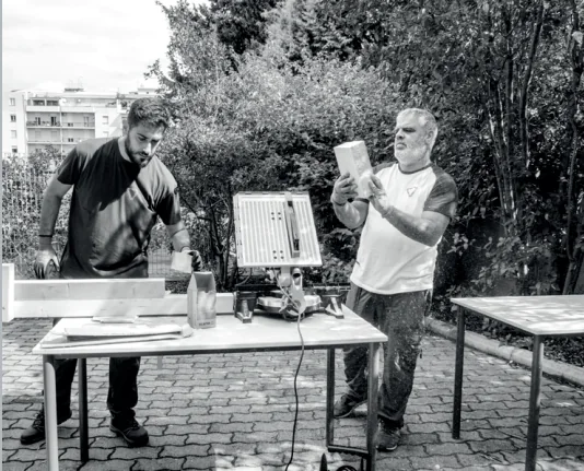 Due partecipanti esaminano disegni e componenti su un tavolo di lavoro all'aperto.