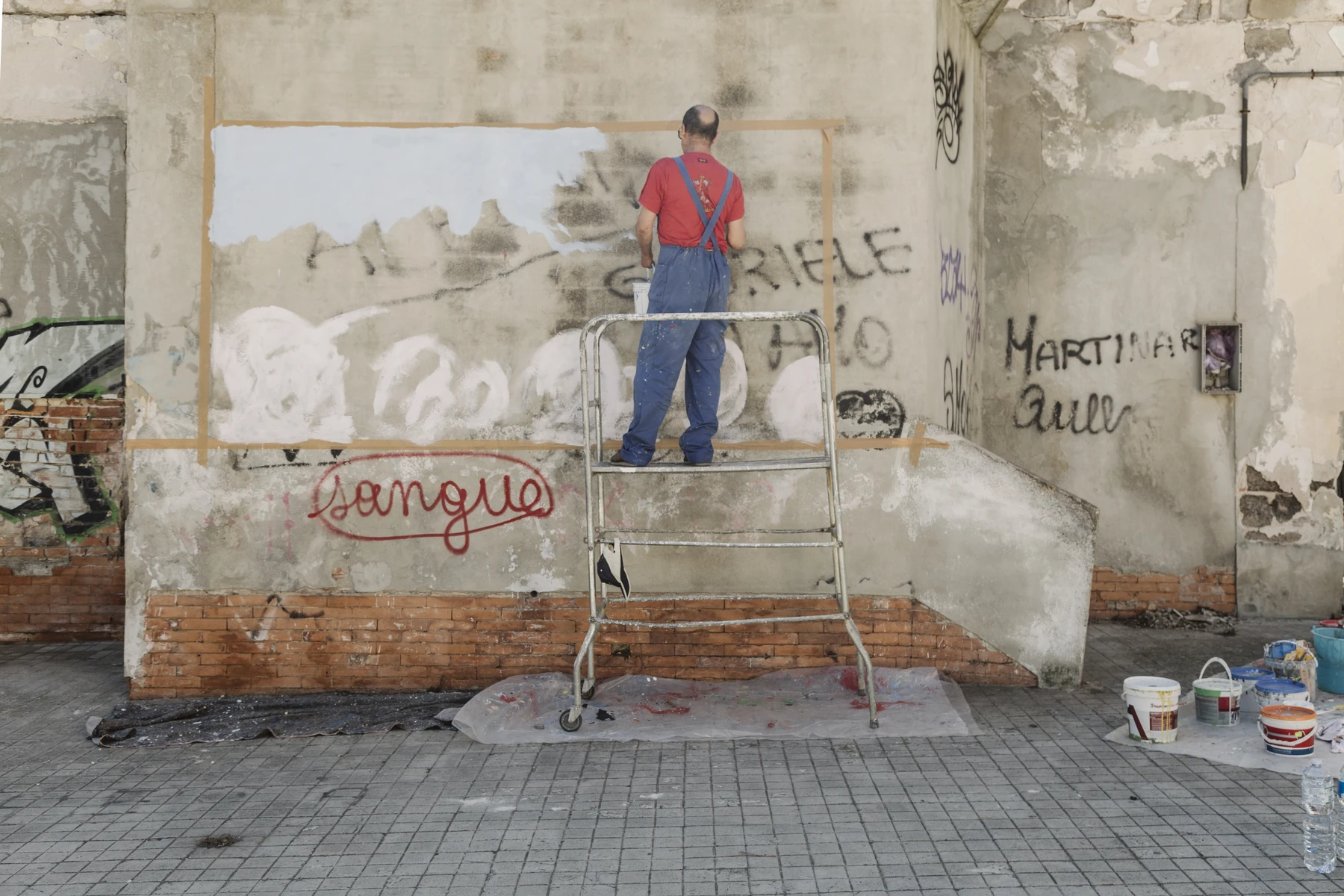 A person on a ladder paints a large pale field on a rough wall covered with writing.